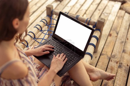 Téléchargez les photos : Femme pigiste assise sur un transat, travaillant à distance depuis la plage, tenant un ordinateur portable sur ses genoux et tapant, copiant de l'espace sur l'écran de l'ordinateur - en image libre de droit