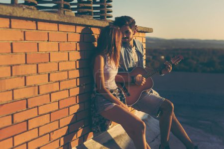 Téléchargez les photos : Couple amoureux traînant à une fête sur le toit, appuyé contre un mur de briques ; gars jouant de la guitare et chantant - en image libre de droit