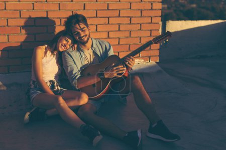 Photo for Couple in love hanging out at a rooftop party, sitting on the floor, leaning against a brick wall and enjoying a beautiful summer sunset - Royalty Free Image