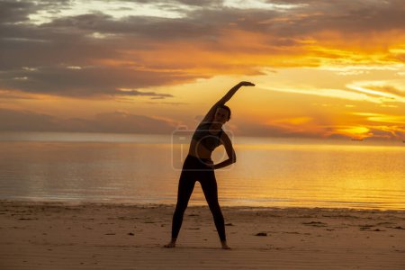 Téléchargez les photos : Femme en forme active s'étendant sur la plage au lever du soleil ; jeune femme travaillant sur la plage tôt le matin - en image libre de droit