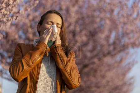 Photo for Portrait of a young woman sneezing and blowing nose outdoor, having trouble with spring pollen allergies and inhaler allergens - Royalty Free Image
