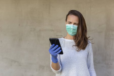 Photo for Young woman wearing medical face protection mask and gloves as part of covid-19 pandemic prevention and protection campaign, typing a text message using smart phone - Royalty Free Image
