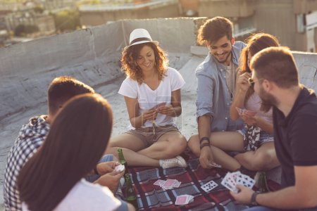 Foto de Grupo de jóvenes sentados en una manta de picnic, divirtiéndose mientras juegan a las cartas en la azotea del edificio. Concéntrate en la chica del medio - Imagen libre de derechos
