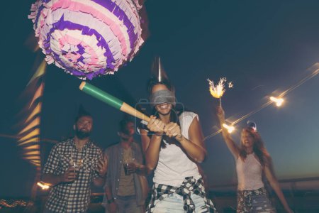 Birthday girl hitting the pinata with baseball bat while her friends are cheering and laughing. Young people having fun at a rooftop birthday party