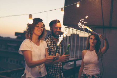 Téléchargez les photos : Groupe de jeunes amis s'amusant à une fête sur le toit, saluant avec des étincelles - en image libre de droit