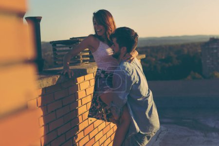 Photo for Couple in love having fun on a building rooftop, enjoying a beautiful summer sunset over the city; guy lifting the girl - Royalty Free Image