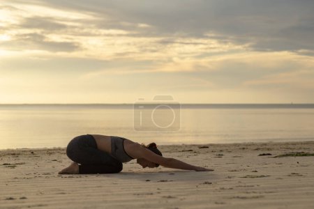 Photo for Young woman training on the beach early in the morning, practising yoga and stretching out, holding Balasana or child's pose - Royalty Free Image