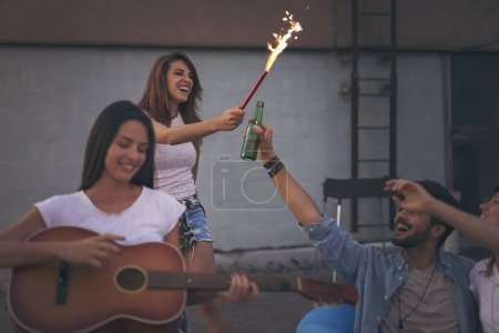 Téléchargez les photos : De jeunes amis s'amusent lors d'une fête estivale sur le toit, jouant de la guitare, chantant, dansant et se relaxant. Concentrez-vous sur la fille agitant avec un scintilleur - en image libre de droit