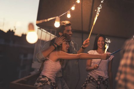 Foto de Grupo de jóvenes amigos divirtiéndose en una fiesta en la azotea, cantando, bailando y saludando con bengalas - Imagen libre de derechos