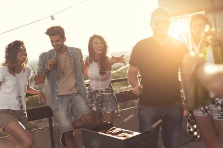 Photo for Young friends making barbecue, drinking beer and enjoying hot summer days having fun on a rooftop party. Focus on the girl in the middle - Royalty Free Image