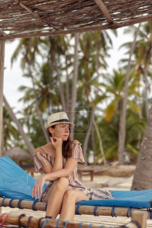 Téléchargez les photos : Belle jeune femme relaxante dans une station balnéaire exotique tropicale, assise sur un transat et profitant de journées d'été ensoleillées pendant ses vacances - en image libre de droit