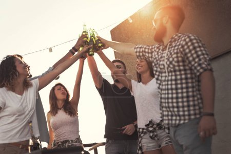 Téléchargez les photos : Groupe de jeunes amis s'amuser à la fête sur le toit, faire un toast et profiter des chaudes journées d'été. Concentrez-vous sur le gars au milieu - en image libre de droit