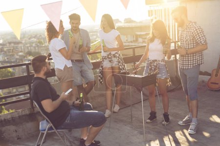Téléchargez les photos : Groupe de jeunes amis s'amuser à la fête sur le toit, faire un barbecue, boire de la bière et profiter des chaudes journées d'été. Concentrez-vous sur les gens à côté de la clôture - en image libre de droit