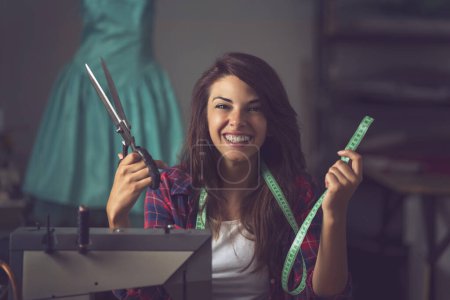 Photo for Young tailor sitting at a sewing machine, holding scissors and a measuring meter and having fun while working in her workshop - Royalty Free Image