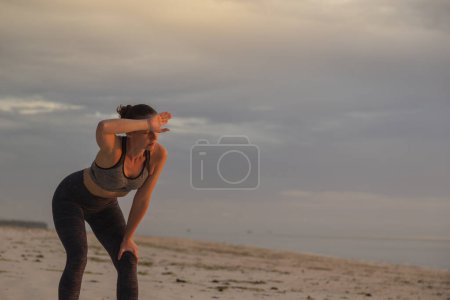 Téléchargez les photos : Jeune femme debout sur la plage après un entraînement intense, s'appuyant sur ses genoux et essuyant la sueur après avoir fait du jogging sur la plage tôt le matin - en image libre de droit