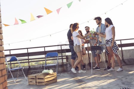 Foto de Grupo de jóvenes amigos divirtiéndose en la fiesta en la azotea, haciendo barbacoa, bebiendo cerveza y disfrutando de los calurosos días de verano. Concéntrate en la pareja en el medio - Imagen libre de derechos