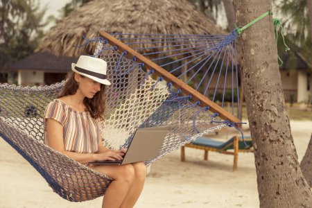 Photo for Female freelancer sitting in a hammock tied up between palm trees on beautiful exotic tropical beach, working on a laptop computer; digital nomad telecommuting - Royalty Free Image