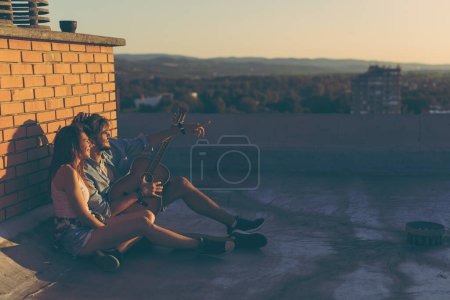 Téléchargez les photos : Couple amoureux traînant à une fête sur le toit, assis sur le sol, appuyé contre un mur de briques et profitant d'un magnifique coucher de soleil d'été - en image libre de droit