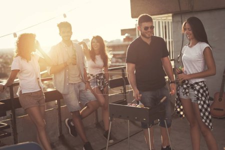Photo for Group of young friends having fun at rooftop party, making barbecue and enjoying hot summer days. Focus on the couple next to the barbecue - Royalty Free Image