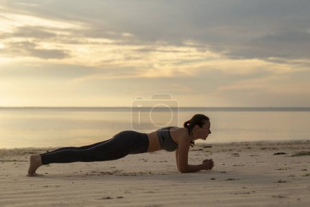 Téléchargez les photos : Vue latérale de la jeune femme travaillant sur la plage le matin, tenant une position de planche avant-bras - en image libre de droit
