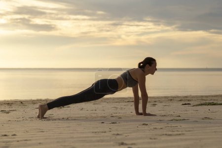 Téléchargez les photos : Vue latérale de la jeune femme travaillant sur la plage le matin, tenant une position de planche à bras droit - en image libre de droit