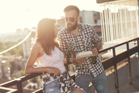 Photo for Young couple having fun at a rooftop party, drinking beer, eating barbecue and chatting - Royalty Free Image