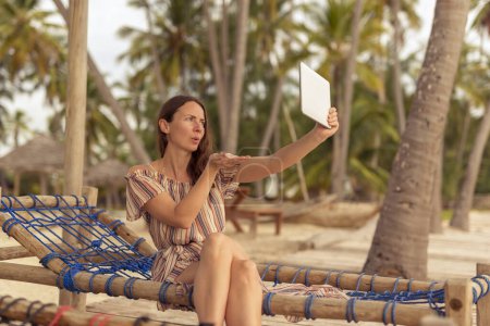 Photo for Woman sitting on a sun bed on a beautiful exotic tropical beach with palm trees, having a video call using a tablet computer, sending kisses - Royalty Free Image