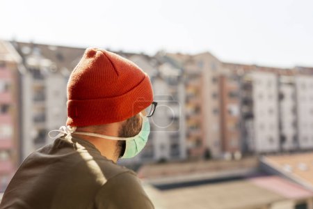 Foto de Retrato de un hombre con máscara médica de protección facial mientras estaba de pie en un balcón en aislamiento domiciliario durante el brote de virus covid-19 - Imagen libre de derechos