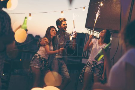 Téléchargez les photos : Groupe de jeunes amis s'amusant à une fête sur le toit, chantant, dansant et saluant avec des étincelles - en image libre de droit
