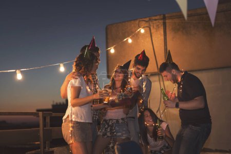 Téléchargez les photos : Groupe de jeunes amis organisant une fête d'anniversaire sur le toit d'un immeuble, chantant une chanson et soufflant une bougie. Focus sur la fille anniversaire - en image libre de droit