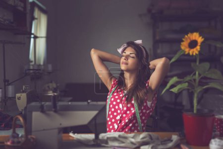 Téléchargez les photos : Belle jeune couturière assise et faisant une pause dans son atelier, détendue et joyeuse - en image libre de droit