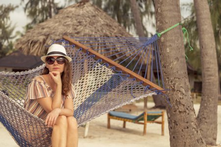 Foto de Mujer joven relajándose mientras está de vacaciones sentado en una hamaca atada entre palmeras en la hermosa playa tropical exótica - Imagen libre de derechos