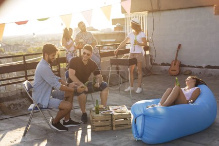 Téléchargez les photos : Groupe de jeunes amis s'amuser à la fête sur le toit, faire un barbecue, boire de la bière et profiter des chaudes journées d'été. Focus sur l'homme en T-shirt noir - en image libre de droit