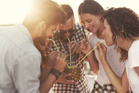 Téléchargez les photos : Groupe de jeunes amis qui s'amusent à la fête sur le toit, en buvant un cocktail d'ananas. Concentrez-vous sur le couple au milieu - en image libre de droit