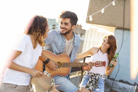 Photo for Three young friends having fun at rooftop party, playing guitar and singing. Focus on the man playing the guitar - Royalty Free Image