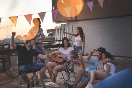 Téléchargez les photos : De jeunes amis s'amusent à une fête sur le toit, jouent de la guitare, chantent, dansent et se relaxent. Concentre-toi sur la fille qui joue de la guitare - en image libre de droit