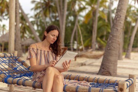 Photo for Female freelancer working on the beach while traveling; digital nomad sitting on a sunbed on a beautiful exotic tropical beach with palm trees, using a tablet computer - Royalty Free Image