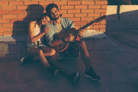 Téléchargez les photos : Couple amoureux traînant à une fête sur le toit, assis sur le sol, appuyé contre un mur de briques et profitant d'un magnifique coucher de soleil d'été - en image libre de droit