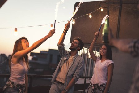 Téléchargez les photos : Groupe de jeunes amis s'amusant à une fête sur le toit, chantant, dansant et saluant avec des étincelles. Concentrez-vous sur le couple à droite - en image libre de droit