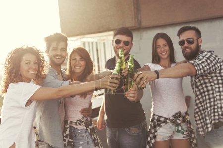 Foto de Grupo de jóvenes amigos divirtiéndose en la fiesta en la azotea, haciendo un brindis con botellas de cerveza y disfrutando de los calurosos días de verano. Concéntrate en la cerveza y la chica de la izquierda. - Imagen libre de derechos