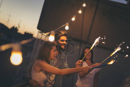 Photo for Group of young friends having fun at a rooftop party, singing, dancing and waving with sparklers. Focus on the guy - Royalty Free Image