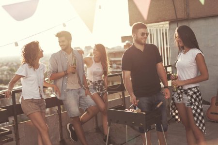 Foto de Grupo de jóvenes amigos divirtiéndose en la fiesta en la azotea, haciendo barbacoa, bebiendo cerveza y disfrutando de los calurosos días de verano. Concéntrate en el hombre al lado de una barbacoa - Imagen libre de derechos