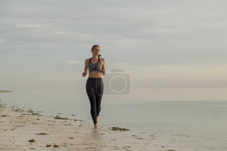 Téléchargez les photos : Jeune femme travaillant sur la plage le matin, faisant du jogging le long de la côte - en image libre de droit
