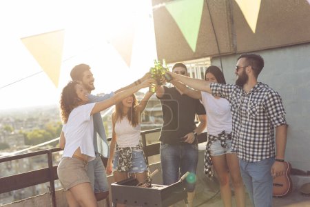 Photo for Group of young friends having fun at rooftop party, making a toast and enjoying hot summer days. Focus on the people on both sides - Royalty Free Image