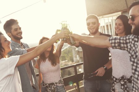 Téléchargez les photos : Groupe de jeunes amis s'amuser à la fête sur le toit, faire un toast avec de la bière et profiter des chaudes journées d'été. Concentrez-vous sur le gars au milieu - en image libre de droit