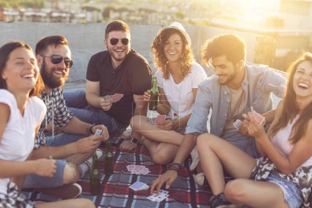 Photo for Group of young people sitting on a picnic blanket, having fun while playing cards on the rooftop. Focus on the couple in the middle - Royalty Free Image