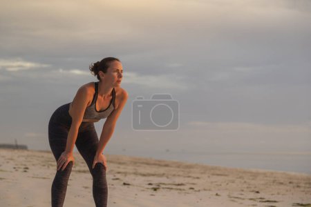 Photo for Young woman standing on the beach after jogging, leaning on her knees and taking a break - Royalty Free Image