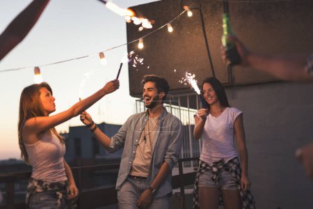 Téléchargez les photos : Groupe de jeunes amis s'amusant à une fête sur le toit, chantant, dansant et saluant avec des étincelles. Concentrez-vous sur le couple à droite - en image libre de droit