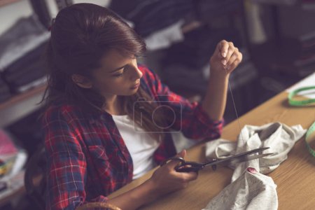 Téléchargez les photos : Jeune couturière faisant quelques réparations, cousant manuellement des boutons sur une chemise et coupant le fil supplémentaire. - en image libre de droit