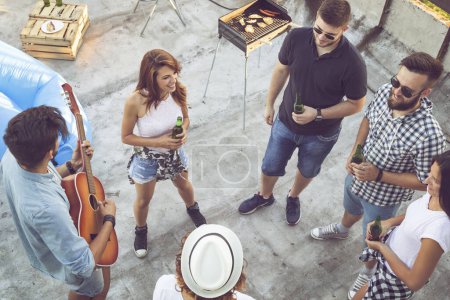 Téléchargez les photos : Vue de dessus d'un groupe de jeunes amis s'amusant à une fête sur le toit, jouant de la guitare, chantant et profitant des chaudes journées d'été. Concentrez-vous sur les gars à droite - en image libre de droit